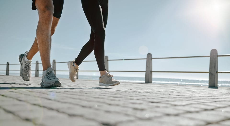 People, legs and running at the beach for exercise, cardio workout or training together outdoors. Leg of friends taking run, walk or jog on warm sunny day by the ocean coast for healthy wellness.
